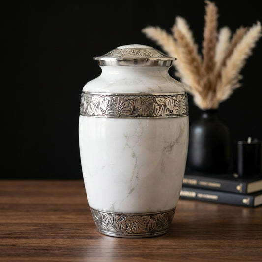A white marble-look cremation urn with ornate silver-toned metal bands and a matching lid, displayed on a wooden surface against a dark background with dried grass and books in the rear.