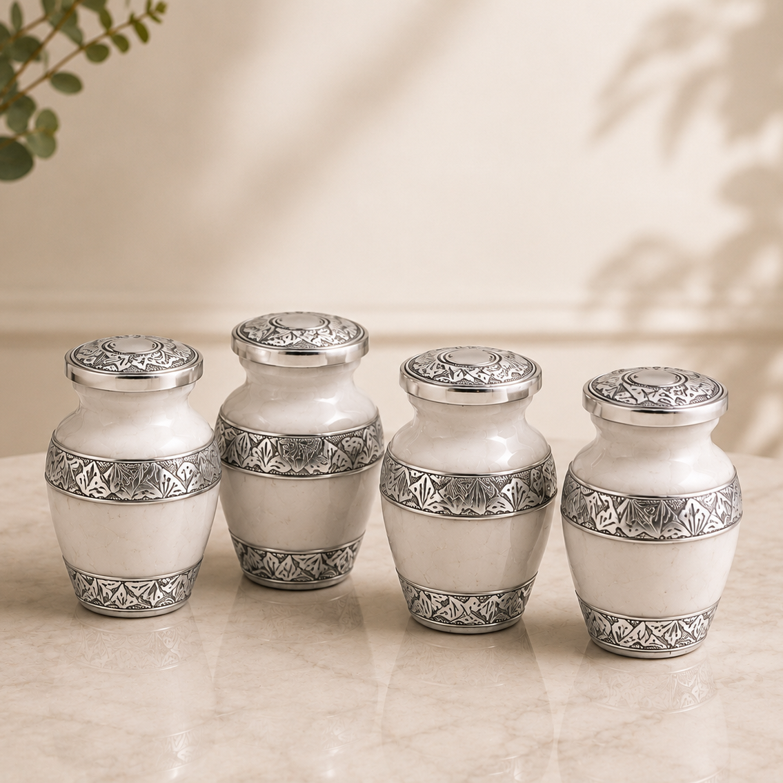 Four small white ceramic vases with ornate silver metal bands and lids, arranged on a light marble surface against a warm beige background.

