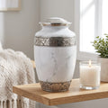 A white marble-look urn with ornate silver-toned metal bands and a matching lid, displayed on a wooden shelf beside a lit candle and a small potted plant in a softly lit room.