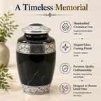 Black cremation urn with silver detailing on a light wooden surface, surrounded by greenery.