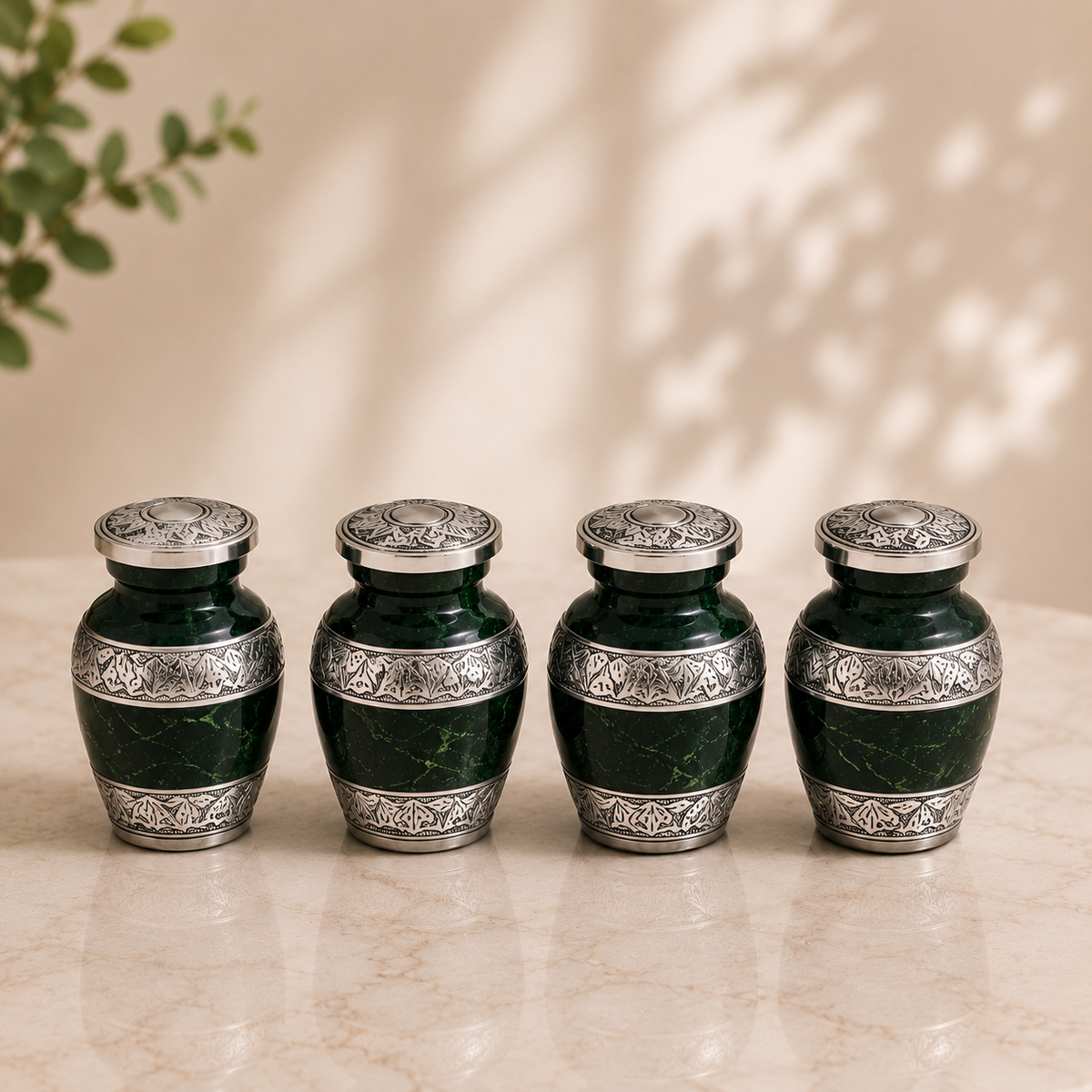 Four small dark green marble urns with ornate silver metal bands and matching silver lids, arranged in a row on a light beige marble surface against a warm beige background with soft leaf shadows.