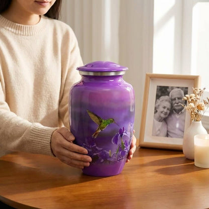 Person holding a purple urn with a hummingbird design on a wooden table.