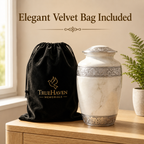 A black velvet drawstring bag with gold TrueHaven Memorials branding sits beside a white marble-style cremation urn with silver decorative bands, displayed on a wooden table in a softly lit interior with a plant in the background.
